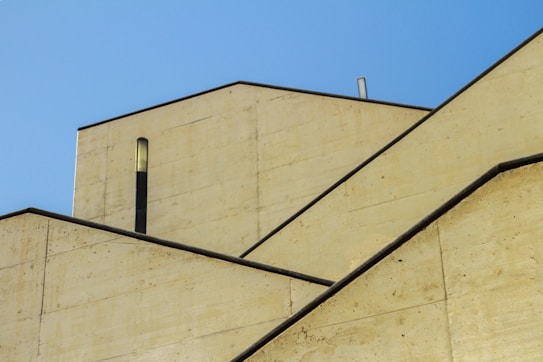 Angular, geometric architectural structures with intersecting lines and muted textures, set against a clear blue sky. The design is minimalist, featuring sharp edges and a modern aesthetic.