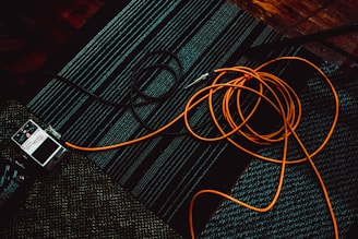 A coiled orange cable and a guitar effects pedal are placed on a dark, patterned carpet. The cable sprawls casually across the floor with visible texture on the carpet.