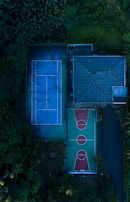 Aerial view of a sports complex featuring a blue tennis court and a green and red basketball court, surrounded by lush greenery and adjacent to a building with a sloped roof.
