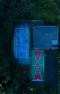 Aerial view of a sports complex featuring a blue tennis court and a green and red basketball court, surrounded by lush greenery and adjacent to a building with a sloped roof.