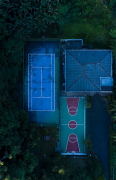 Aerial view of a sports complex featuring a blue tennis court and a green and red basketball court, surrounded by lush greenery and adjacent to a building with a sloped roof.