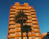 A tall apartment building with a brown and yellow facade stands against a clear blue sky. In front of the building, a large palm tree casts a shadow across the structure. The building has multiple balconies and windows, with a 'for sale' sign displayed on one of the balconies.