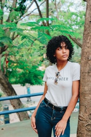 A person with curly hair wearing a striped shirt with the word 'INFLUENCER' on it stands next to a tree. The scene is set outdoors with lush green foliage in the background. The person appears contemplative, looking off to the side.
