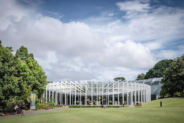 The Calyx, a modern glass-roofed pavilion in Sydney's Royal Botanic Garden, stands on a lawn under a partly cloudy sky.