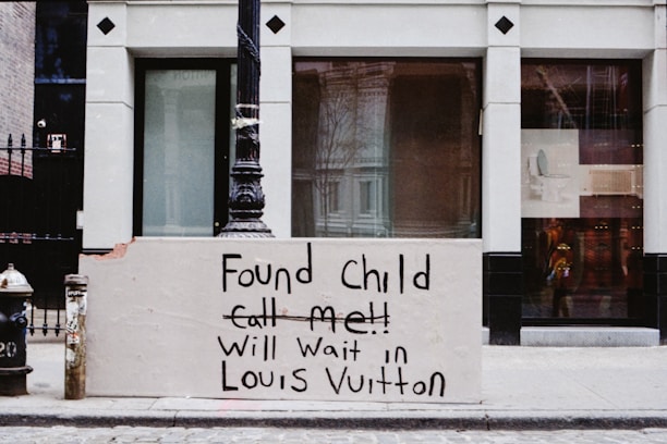 A signboard on a city sidewalk has a handwritten message stating that a child has been found and the writer will wait in a Louis Vuitton store. The sign is placed against the base of a lamppost, next to a fire hydrant. The background features a storefront with large windows and some reflections, typical of a commercial area.