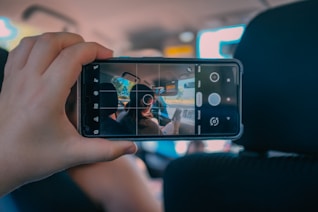 A hand is holding a smartphone in a car, with the screen showing the camera interface. The camera is pointed toward the interior of the car where two people are sitting in the front seats. The focus on the phone screen contrasts with the slightly blurred car background.