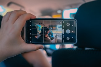 A hand is holding a smartphone in a car, with the screen showing the camera interface. The camera is pointed toward the interior of the car where two people are sitting in the front seats. The focus on the phone screen contrasts with the slightly blurred car background.