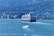 A large cruise ship sails through a wide body of water, surrounded by several smaller boats. In the background, there is a cityscape with numerous buildings and a dense forest area further behind. A bridge spans across the scene, adding a dynamic element to the view.