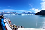A group of people stand on the deck of a cruise ship, observing a vast blue glacier across the calm waters. Snow-capped mountains rise majestically in the background under a clear blue sky.
