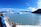 A group of people stand on the deck of a cruise ship, observing a vast blue glacier across the calm waters. Snow-capped mountains rise majestically in the background under a clear blue sky.