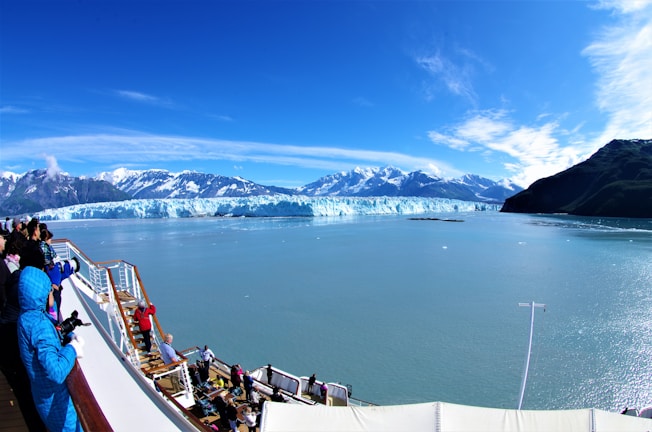 A group of people stand on the deck of a cruise ship, observing a vast blue glacier across the calm waters. Snow-capped mountains rise majestically in the background under a clear blue sky.