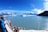 A group of people stand on the deck of a cruise ship, observing a vast blue glacier across the calm waters. Snow-capped mountains rise majestically in the background under a clear blue sky.