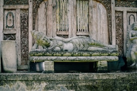 A detailed stone sculpture of a reclining figure, possibly a deity, is set against an intricately carved wooden backdrop. The serene expression and detailed design suggest historical or cultural significance, surrounded by ornate wooden architecture.