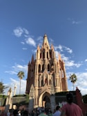 A large, ornate Gothic-style church with spires reaching towards a bright blue sky. The facade is a pinkish hue, typical of neo-Gothic architecture. Palm trees and people are visible around the structure, which is surrounded by an iron fence.