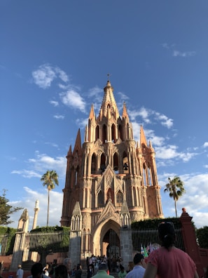 A large, ornate Gothic-style church with spires reaching towards a bright blue sky. The facade is a pinkish hue, typical of neo-Gothic architecture. Palm trees and people are visible around the structure, which is surrounded by an iron fence.
