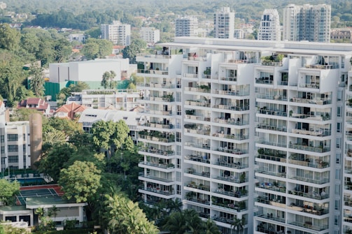 High-rise residential buildings dominate the scene, displaying numerous balconies filled with plants and outdoor furniture. The surrounding area includes lush greenery, a tennis court, and a mix of smaller buildings. In the background, tall skyscrapers and a wooded area are visible, suggesting a blend of urban and natural landscapes.