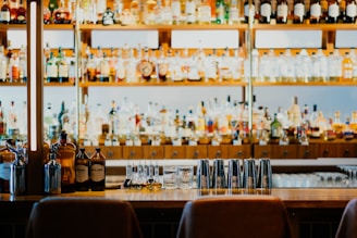 A well-stocked bar features shelves filled with various bottles of liquor and spirits. In the foreground, there is a counter with several metal cocktail shakers, small bottles, and a glass. The lighting and arrangement create a welcoming and organized atmosphere.