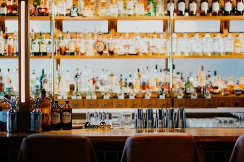 A well-stocked bar features shelves filled with various bottles of liquor and spirits. In the foreground, there is a counter with several metal cocktail shakers, small bottles, and a glass. The lighting and arrangement create a welcoming and organized atmosphere.