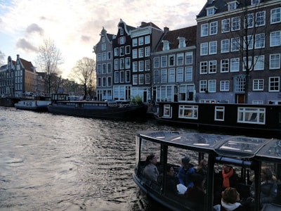 A picturesque canal scene with traditional narrow buildings alongside the water. Several houseboats are moored along the bank, and a glass-roofed tour boat filled with people is passing by. The sky is partly cloudy, with a warm glow from the setting or rising sun.