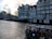 A picturesque canal scene with traditional narrow buildings alongside the water. Several houseboats are moored along the bank, and a glass-roofed tour boat filled with people is passing by. The sky is partly cloudy, with a warm glow from the setting or rising sun.