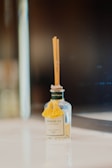 A small, clear glass bottle with a decorative yellow tassel on the front. It contains several thin reed sticks emerging from the top, indicating it is a reed diffuser. The bottle has a label with the brand name 'Shangri-La' and is placed on a reflective, smooth surface against a blurred dark background.
