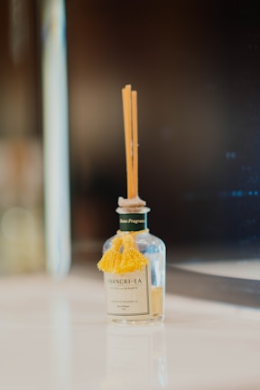 A small, clear glass bottle with a decorative yellow tassel on the front. It contains several thin reed sticks emerging from the top, indicating it is a reed diffuser. The bottle has a label with the brand name 'Shangri-La' and is placed on a reflective, smooth surface against a blurred dark background.