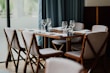A neatly set dining table with six chairs surrounding it. White napkins are placed on the wooden table along with empty wine glasses. The chairs have wooden frames and light-colored cushions. The background features a large window with soft curtains, bringing in natural light.
