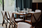 A neatly set dining table with six chairs surrounding it. White napkins are placed on the wooden table along with empty wine glasses. The chairs have wooden frames and light-colored cushions. The background features a large window with soft curtains, bringing in natural light.