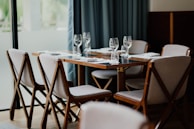 A neatly set dining table with six chairs surrounding it. White napkins are placed on the wooden table along with empty wine glasses. The chairs have wooden frames and light-colored cushions. The background features a large window with soft curtains, bringing in natural light.