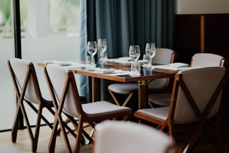 A neatly set dining table with six chairs surrounding it. White napkins are placed on the wooden table along with empty wine glasses. The chairs have wooden frames and light-colored cushions. The background features a large window with soft curtains, bringing in natural light.
