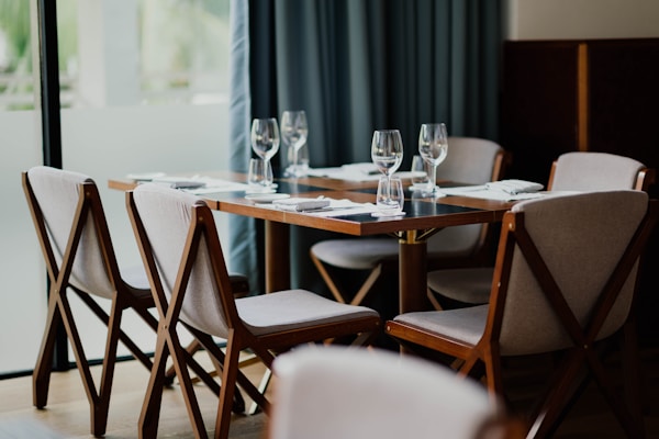 A neatly set dining table with six chairs surrounding it. White napkins are placed on the wooden table along with empty wine glasses. The chairs have wooden frames and light-colored cushions. The background features a large window with soft curtains, bringing in natural light.