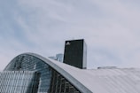 A modern architectural structure with curved, glass-covered surfaces dominates the foreground, while a tall, black skyscraper labeled 'AREVA' rises in the background. The sky is overcast with soft, diffused clouds.