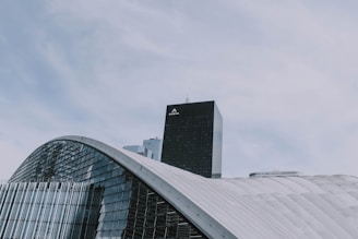 A modern architectural structure with curved, glass-covered surfaces dominates the foreground, while a tall, black skyscraper labeled 'AREVA' rises in the background. The sky is overcast with soft, diffused clouds.