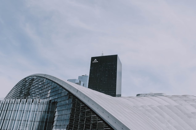 A modern architectural structure with curved, glass-covered surfaces dominates the foreground, while a tall, black skyscraper labeled 'AREVA' rises in the background. The sky is overcast with soft, diffused clouds.