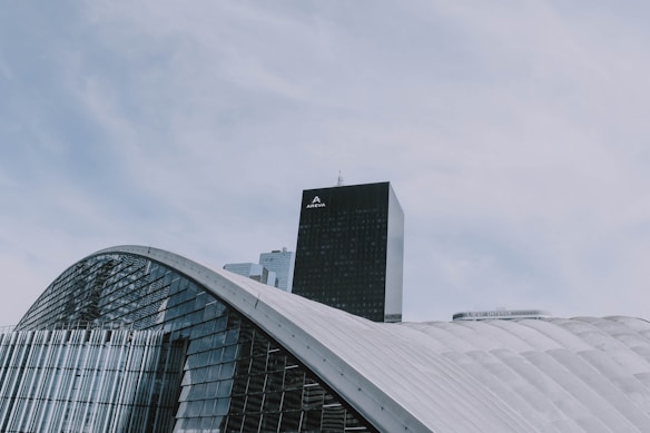 A modern architectural structure with curved, glass-covered surfaces dominates the foreground, while a tall, black skyscraper labeled 'AREVA' rises in the background. The sky is overcast with soft, diffused clouds.