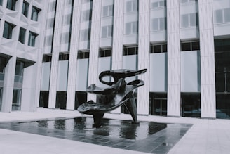 A large, abstract black sculpture is situated in a shallow reflecting pool in front of a modern building with geometric patterns on the facade. The sculpture features smooth, curved shapes and appears to be made of metal.