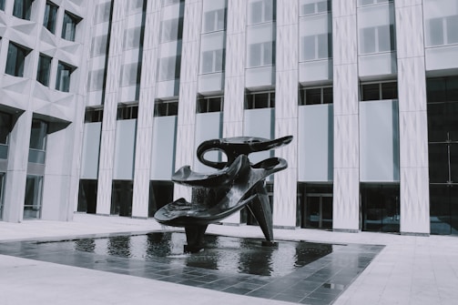 A large, abstract black sculpture is situated in a shallow reflecting pool in front of a modern building with geometric patterns on the facade. The sculpture features smooth, curved shapes and appears to be made of metal.