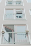 A facade of a building with two rows of large glass windows and light green metal railings. There are small potted plants attached to the railings, adding a touch of greenery. The overall design is minimalistic with clean lines.
