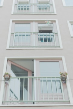 A facade of a building with two rows of large glass windows and light green metal railings. There are small potted plants attached to the railings, adding a touch of greenery. The overall design is minimalistic with clean lines.