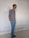 A person stands in a relaxed pose against a plain wall, wearing sunglasses, a striped shirt, blue pants, and black shoes.