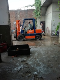 A forklift is parked in a concrete area near a brick wall. Two men are engaged in a conversation near the forklift, and a motorcycle is parked nearby. There is vegetation climbing on the wall and reaching towards an overhanging roof.