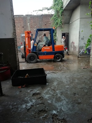 A forklift is parked in a concrete area near a brick wall. Two men are engaged in a conversation near the forklift, and a motorcycle is parked nearby. There is vegetation climbing on the wall and reaching towards an overhanging roof.
