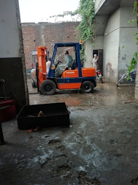 A forklift is parked in a concrete area near a brick wall. Two men are engaged in a conversation near the forklift, and a motorcycle is parked nearby. There is vegetation climbing on the wall and reaching towards an overhanging roof.