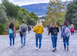 A group of five young people walk down a paved pathway lined with trees, with a mountainous landscape in the background. They all carry backpacks and wear casual clothing, including jeans and jackets. Additional individuals can be seen in the distance.