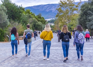 A group of five young people walk down a paved pathway lined with trees, with a mountainous landscape in the background. They all carry backpacks and wear casual clothing, including jeans and jackets. Additional individuals can be seen in the distance.