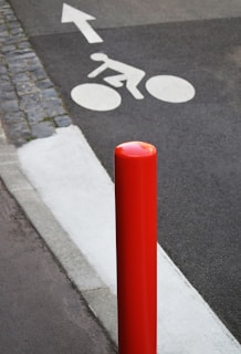 A red bollard stands prominently next to a paved pathway. On the ground, there is a white painted symbol of a cyclist and an arrow indicating the direction. The left side of the path is bordered by cobblestones.