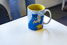 A colorful mug featuring a cartoon character holding several cookies, with a background pattern of repeating text. The interior of the mug is yellow, and it is placed on a white table with a partial view of someone standing nearby.