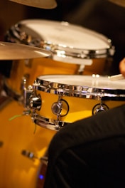 A close-up view of a polished drum set with a prominent metallic snare drum and cymbals. The drums have a glossy, reflective surface, and there is a hand partially visible, suggesting a musician playing.