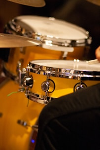 A close-up view of a polished drum set with a prominent metallic snare drum and cymbals. The drums have a glossy, reflective surface, and there is a hand partially visible, suggesting a musician playing.
