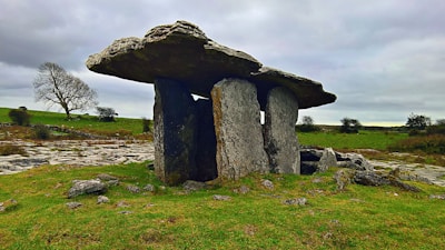 A large ancient stone structure stands in a grassy landscape with cloudy skies. The stone formation consists of two large vertical slabs supporting a massive horizontal stone, resembling a dolmen or megalithic tomb. The surrounding area features patches of grass, several scattered rocks, and a few leafless trees in the distance.